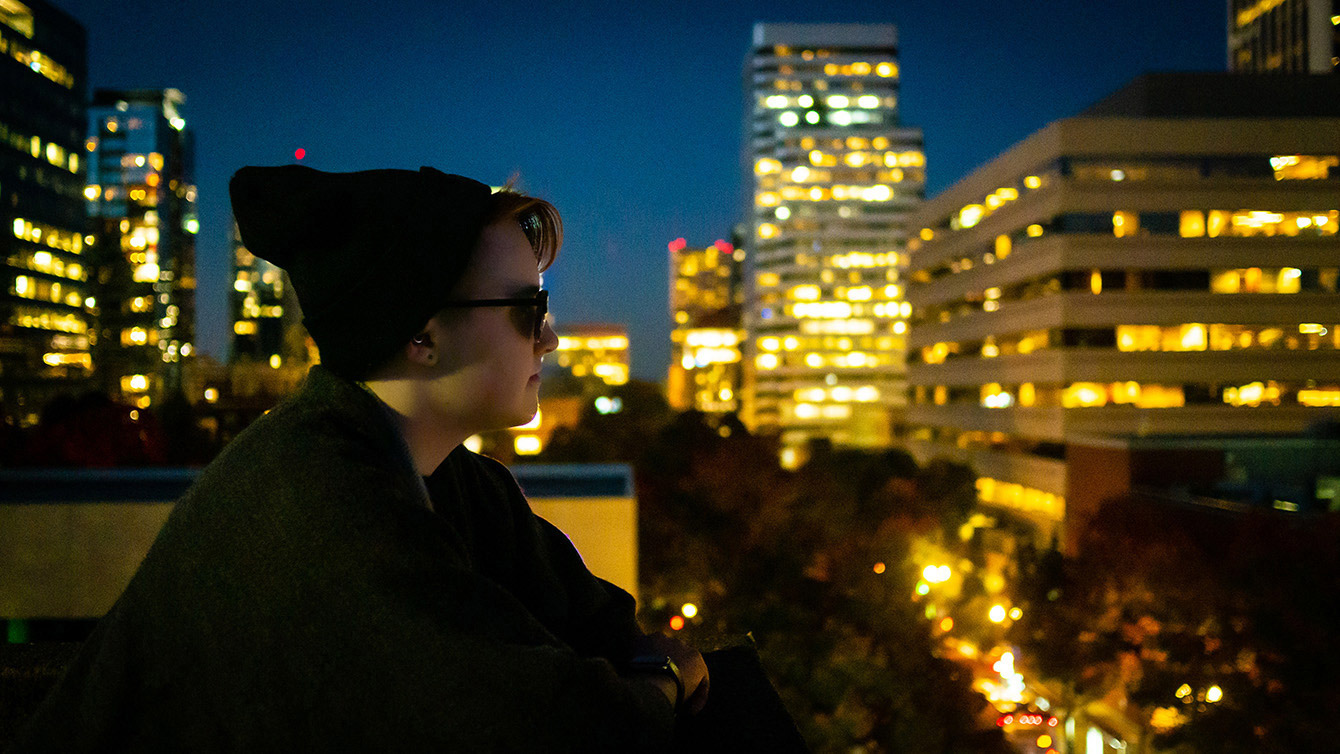 Photo of a man at night with city in background.
